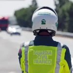 Un agente de Tráfico de la Guardia Civil, con casco y chaleco de alta visibilidad, se encuentra de pie al borde de la carretera, de espaldas a la cámara, controlando a los conductores por exceso de velocidad, con vehículos borrosos y una carretera al fondo.