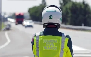 Un agente de Tráfico de la Guardia Civil, con casco y chaleco de alta visibilidad, se encuentra de pie al borde de la carretera, de espaldas a la cámara, controlando a los conductores por exceso de velocidad, con vehículos borrosos y una carretera al fondo.