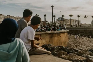 Varias personas están sentadas y de pie en una playa rocosa, con palmeras y edificios al fondo. Tres personas están sentadas en primer plano, mirando hacia la multitud reunida a lo largo de la orilla bajo un cielo nublado: una escena que recuerda a las recientes concentraciones de migrantes en Ceuta, de las que se habla a menudo en las redes sociales.