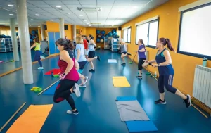 Un grupo de personas vestidas con ropa deportiva hace ejercicio juntas en un gimnasio muy bien iluminado, equipado con colchonetas, espejos y ventanas. Algunas corren sin moverse del sitio, mientras que otras sostienen palos, participando en una clase de entrenamiento en grupo que forma parte de las escuelas deportivas municipales de Rivas. Puedes solicitar plaza para apuntarte y disfrutar de estas dinámicas sesiones con la comunidad.