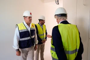 Tres hombres con cascos blancos y chalecos de seguridad amarillos se encuentran dentro de un edificio en construcción, que forma parte de un proyecto de viviendas públicas de 2023 en la Comunidad de Madrid. Dos de ellos están uno frente al otro, mientras que el tercero, en primer plano, habla y gesticula con la mano. El entorno está muy bien iluminado.