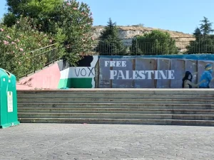 Una escalera de hormigón conduce a un mural en apoyo a Palestina con las palabras «FREE PALESTINE» y la palabra «VOX» sobre un fondo que recuerda a una bandera. A la izquierda de esta escena urbana, que recuerda a los espacios cercanos a Miguel Ríos, se aprecian árboles, un contenedor de reciclaje y referencias a «Convocatoria en Rivas».