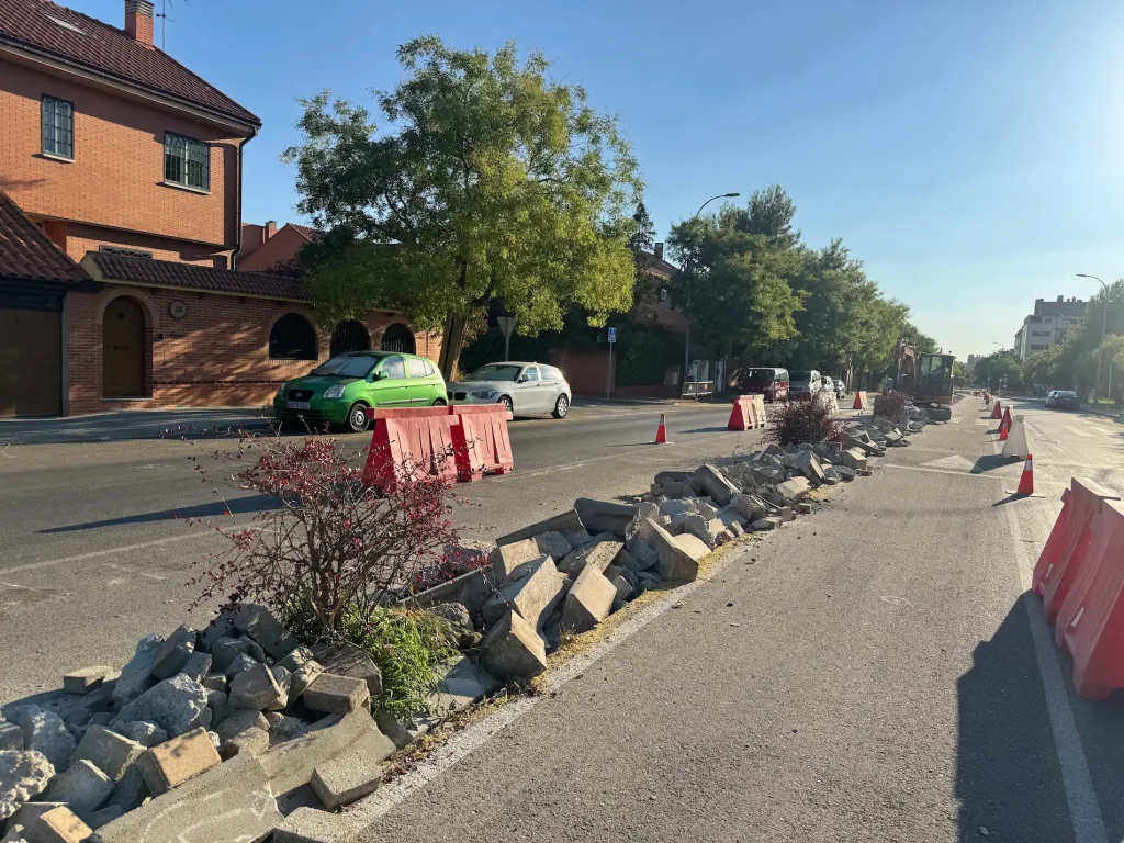 Una calle de Rivas presenta vallas de obra y conos junto a un montón de bloques de hormigón rotos procedentes de una reciente retirada de árboles. Hay coches aparcados a lo largo de la carretera y las casas se alinean a ambos lados de la calle bajo un cielo despejado.