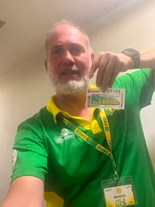 Un hombre con una camiseta verde y amarilla de la ONCE sostiene un boleto de lotería. Lleva barba, lleva una insignia y se encuentra en el interior, posiblemente preparándose para el «Cuponazo ONCE» o el sorteo de los «premios Rivas».