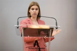 Una mujer con una chaqueta de color coral está de pie en un atril transparente con micrófonos, gesticulando con la mano derecha. En el atril se ven el logotipo y el texto del Ayuntamiento de Alcalá de Henares. El fondo es liso y gris. Judith Piquet, una de las alcaldesas socialistas vinculadas al FMM, se dirige a su público con seguridad.