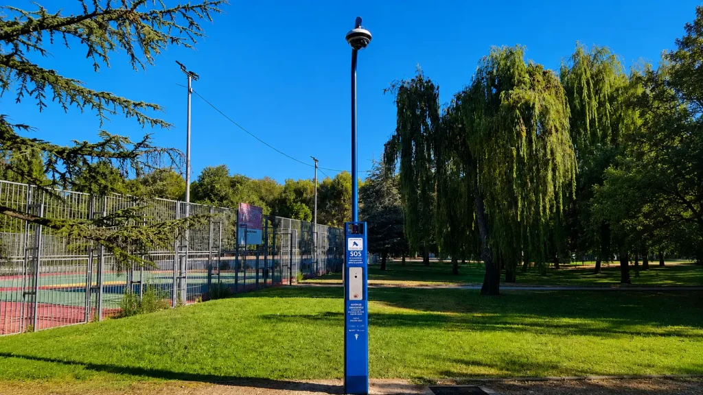 Una cabina azul de llamadas de emergencia, uno de los «SOS postes» de Rivas, equipada con una cámara de seguridad, se encuentra en una zona con césped cerca de una pista de tenis y de unos árboles, en un parque bajo un cielo azul y despejado, lo que aporta mayor seguridad y apoyo frente a la violencia machista.