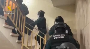 Cuatro agentes de policía, equipados con material táctico y cascos, suben por una escalera. En sus uniformes se lee la inscripción «GUARDIA CIVIL USECIC», lo que indica su participación en operaciones especializadas contra los ladrones de viviendas. La escena se desarrolla en el interior de un edificio en Rivas, con paredes de color beige y un pasamanos amarillo.
