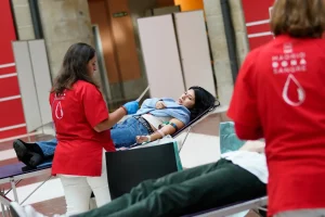 Una mujer dona sangre tumbada en una camilla, atendida por un trabajador que lleva una camiseta roja. En primer plano se ve parcialmente a otro donante. El escenario parece ser una campaña de donación de sangre en un recinto cerrado, que contribuye a mantener las reservas de sangre vitales para la Comunidad de Madrid.