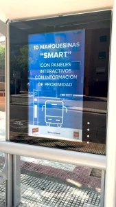 Un cartel en una parada de autobús, redactado en español, anuncia 10 marquesinas inteligentes con paneles interactivos que ofrecen información de proximidad e incluyen mapas interactivos. El cartel incluye iconos de un autobús y un punto de información, mientras que se aprecian los edificios cercanos y la acera de Rivas.