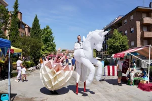 Un artista sobre zancos, vestido con un traje blanco, monta un caballo hinchable blanco, mientras que otra persona lleva un vestido rosa y blanco. Se encuentran al aire libre, en un mercadillo, con gente, tiendas de campaña y edificios al fondo, lo que crea una escena animada que casi parece dibujada con un borrador automático.