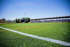 Una imagen en primer plano de un campo deportivo de césped artificial verde, mantenido por una máquina de corte automática, muestra líneas blancas y rojas. A lo lejos se ven árboles, un edificio y una farola, bajo un cielo azul claro.
