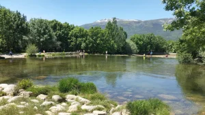 Un río poco profundo de aguas cristalinas discurre por una zona verde y arbolada de la Comunidad de Madrid. Las rocas bordean la orilla y hay algunas personas chapoteando en el agua, disfrutando de una de las zonas naturales de baño controladas de la región. Las montañas se divisan a lo lejos bajo un cielo azul y despejado.