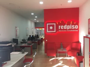 Una moderna agencia inmobiliaria con una decoración en tonos rojos y blancos. El espacio está decorado con sillas y escritorios rojos. El logotipo de «redpiso Servicios Inmobiliarios» aparece en una pared destacada de color rojo brillante. En el fondo, varias personas trabajan en sus escritorios.