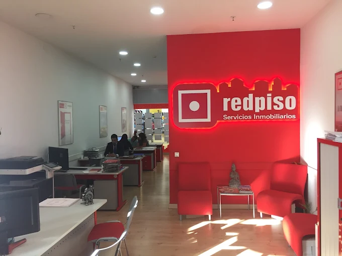 Una moderna agencia inmobiliaria con una decoración en tonos rojos y blancos. El espacio está decorado con sillas y escritorios rojos. El logotipo de «redpiso Servicios Inmobiliarios» aparece en una pared destacada de color rojo brillante. En el fondo, varias personas trabajan en sus escritorios.