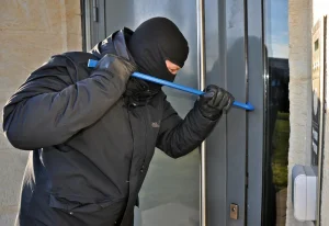 Una persona que lleva una máscara negra, guantes y una chaqueta utiliza una palanca azul para forzar una puerta de cristal, lo que pone de relieve la importancia de reforzar la seguridad para proteger su vivienda en el este de Madrid.