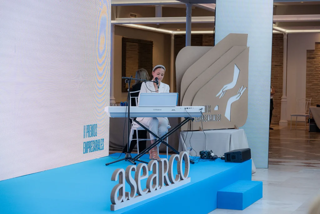 Una persona vestida de blanco está sentada frente a un teclado en un escenario azul, cantando por un micrófono. El escenario cuenta con un cartel de los Premios Empresariales de ASEARCO y un fondo decorativo con manos. La iluminación es intensa y la sala tiene paredes blancas y una decoración moderna.
