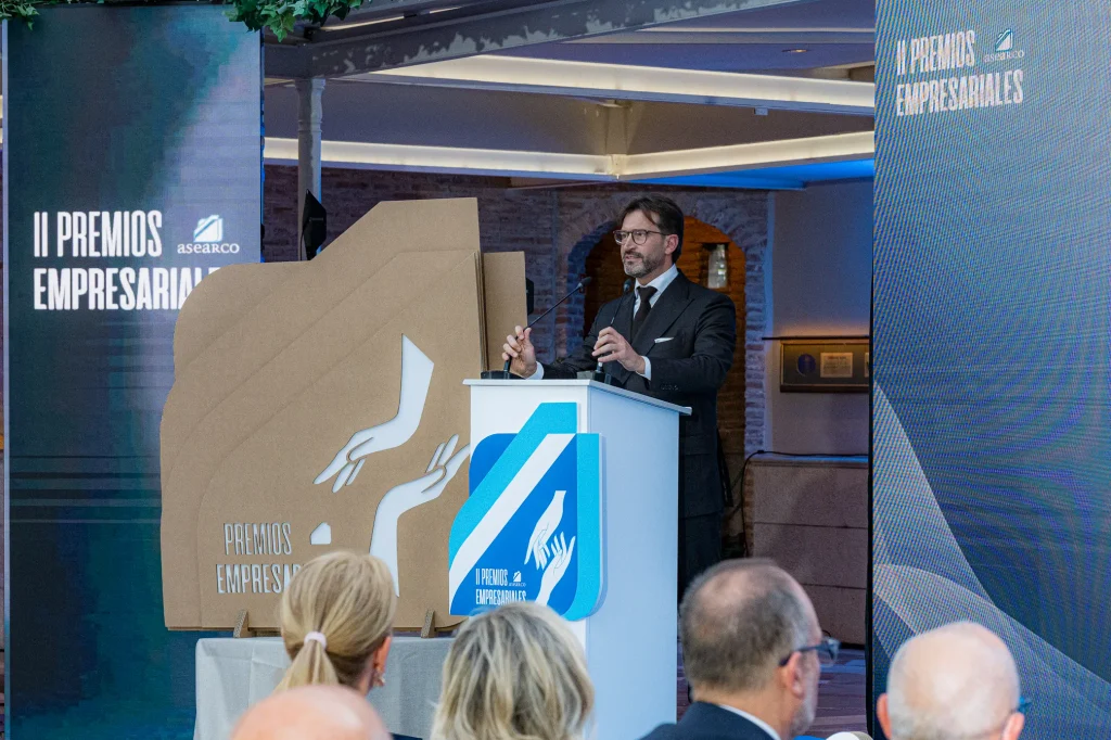 Un hombre vestido con traje habla desde un atril durante la ceremonia de los II Premios Empresariales de ASEARCO. En primer plano hay varias personas sentadas. Al fondo se ven grandes pancartas del evento y una escultura de cartón con dibujos hechos a mano.