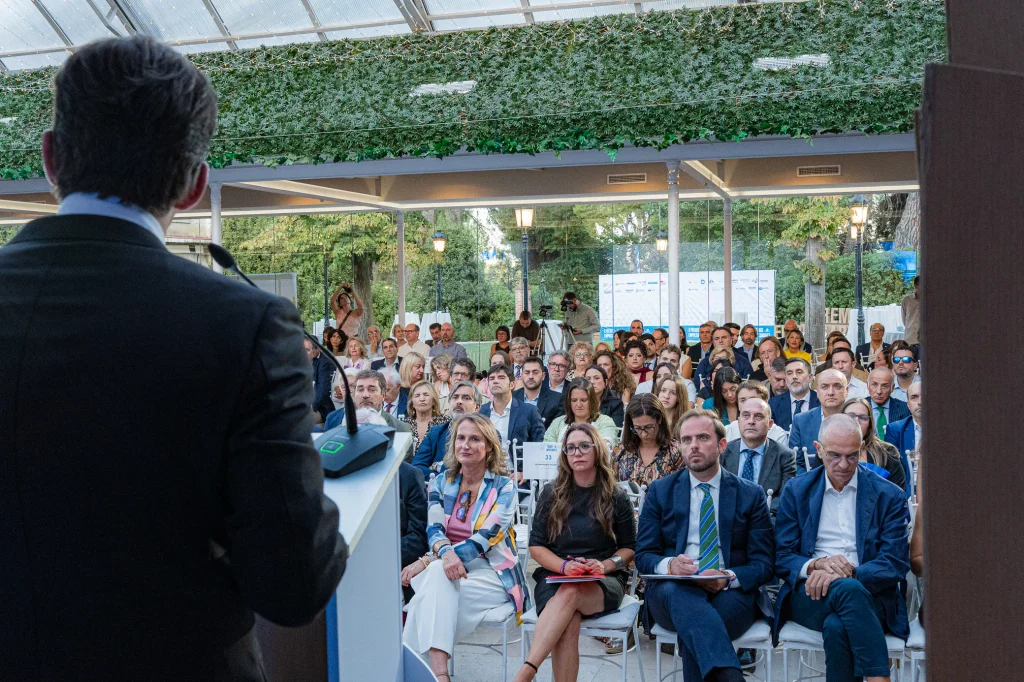 Una persona habla desde un atril ante un público sentado en un recinto luminoso con techo de cristal durante la ceremonia de los Premios Empresariales de ASEARCO. Los asistentes parecen atentos; se trata de un grupo mixto de hombres y mujeres vestidos con traje de negocios. Las paredes superiores del recinto están decoradas con plantas.