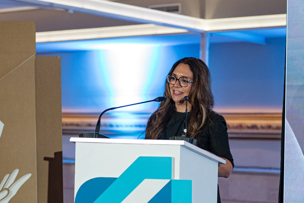Una mujer con gafas y un top negro habla desde un atril blanco con motivos geométricos azules, de pie frente a los micrófonos en un acto celebrado en un recinto cerrado con iluminación azul, durante los Premios Empresariales de ASEARCO.