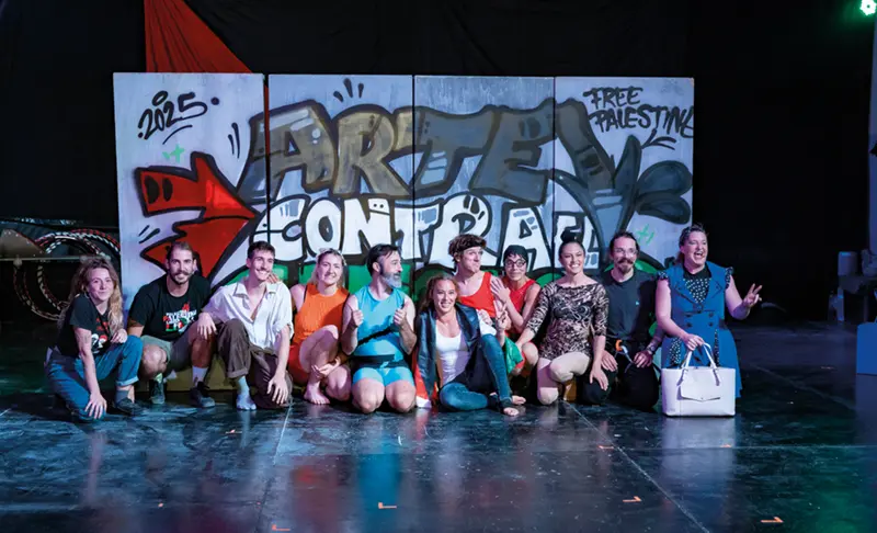 Un grupo de trece personas posa y sonríe frente a una pared cubierta de grafitis en la que se lee «ARTE CONTRA» y «FREE PALESTINE». La mayoría está en cuclillas o de rodillas; el escenario parece ser un interior, en un escenario, lo que sugiere una reunión animada, posiblemente relacionada con Festaclown 2026.