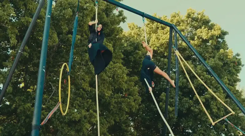 Dos personas realizan acrobacias en cuerdas sujetas a una alta estructura metálica al aire libre durante el Festaclown 2026. Una lleva un traje negro y la otra, uno oscuro. Los árboles verdes llenan el fondo bajo un cielo despejado.