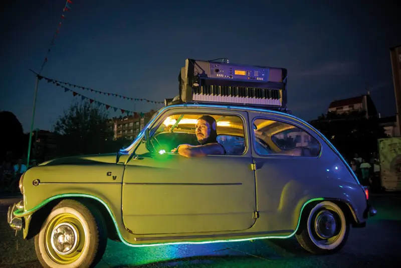 Un hombre está sentado en el interior de un pequeño coche clásico por la noche, iluminado por luces verdes y amarillas, con un teclado y un sintetizador colocados en el techo del coche: una escena llamativa que sin duda será uno de los momentos más destacados del Festaclown 2026.