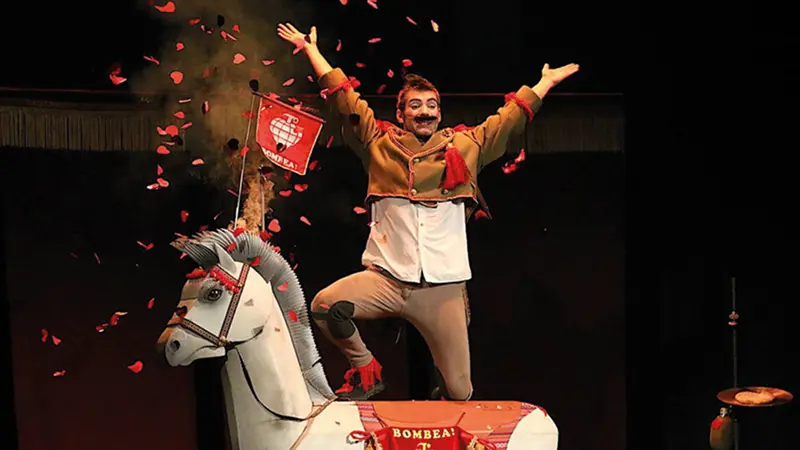 Un artista con bigote posa sobre un caballo de juguete blanco, sosteniendo una bandera roja en Festaclown 2026. A su alrededor cae confeti rojo mientras levanta los brazos y sonríe. El fondo es oscuro y se ven elementos del decorado.