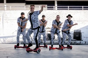 Cinco personas vestidas con trajes a juego en blanco y negro realizan una coreografía sobre hoverboards frente a una pared de ladrillo blanco, con baterías y altavoces visibles al fondo: una escena llena de energía del Festival de Cultura en la Calle 2026.