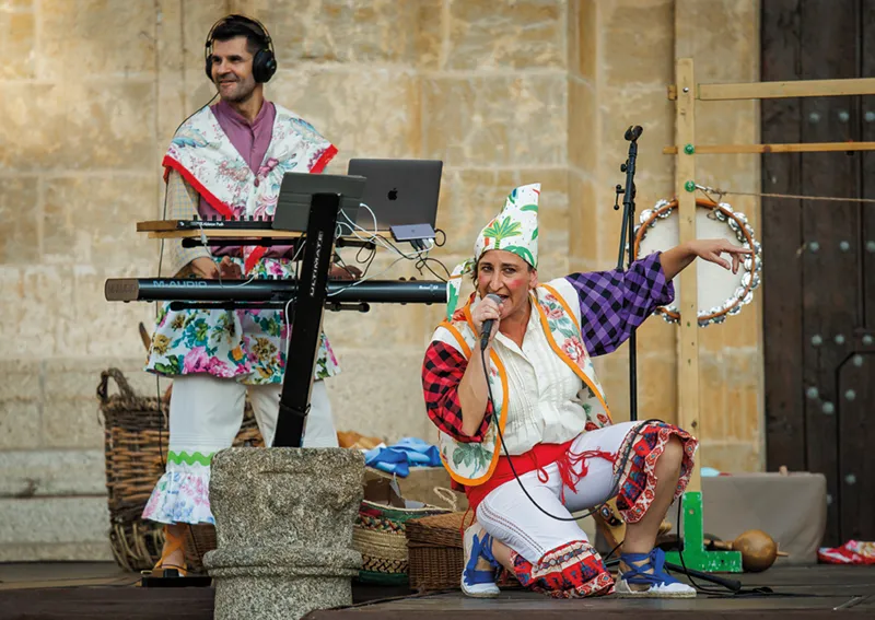 En el Festival de Cultura en la Calle 2026, dos artistas que actúan en el escenario lucen trajes coloridos y eclécticos. Uno toca el teclado y lleva auriculares, mientras que el otro está arrodillado, canta por un micrófono y sostiene una pandereta. El fondo es una pared de piedra.