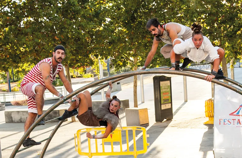 Cuatro artistas posan sobre una estructura metálica en forma de arco y a su alrededor, al aire libre, en el Festival de Cultura en la Calle 2026. Una persona cuelga boca abajo, mientras que los demás están sentados o agachados encima. Al fondo se ven árboles y barreras amarillas.