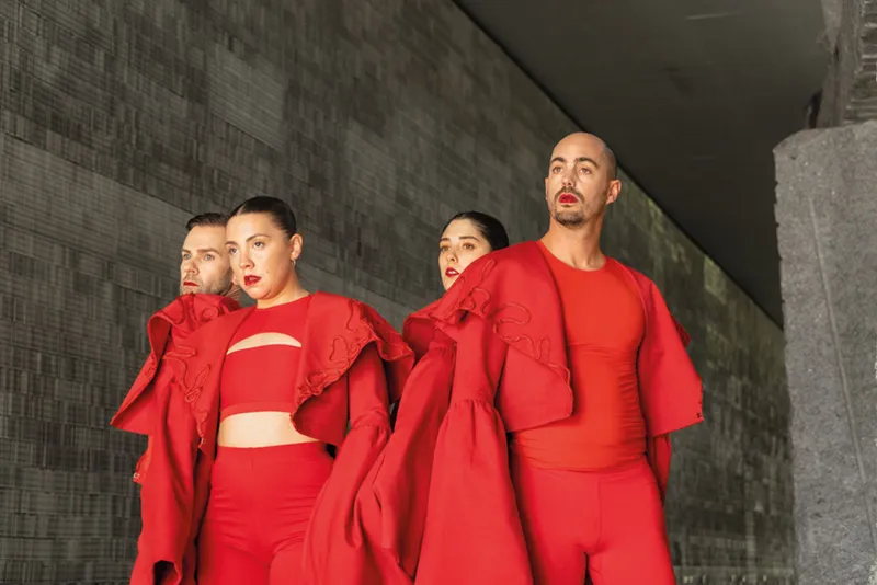 Cuatro personas, de pie una al lado de la otra, visten conjuntos rojos a juego con mangas espectaculares y posan al aire libre frente a una pared gris moderna con textura. Con expresiones serias, miran hacia delante, encarnando el espíritu vibrante del Festival de Cultura en la Calle 2026.