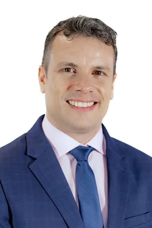 Un hombre con traje azul, camisa rosa claro y corbata azul sonríe a la cámara ante un fondo blanco liso, poco después de que se anunciara la dimisión del concejal del PP de Rivas.