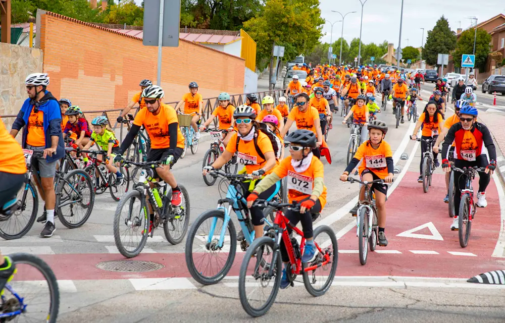 Un numeroso grupo de personas de diversas edades, vestidas con camisetas naranjas y cascos, pedalean juntas en bicicleta por una calle de la ciudad durante la Fiesta de la Bici en Rivas, disfrutando de una de las populares rutas familiares de este animado evento ciclista diurno.