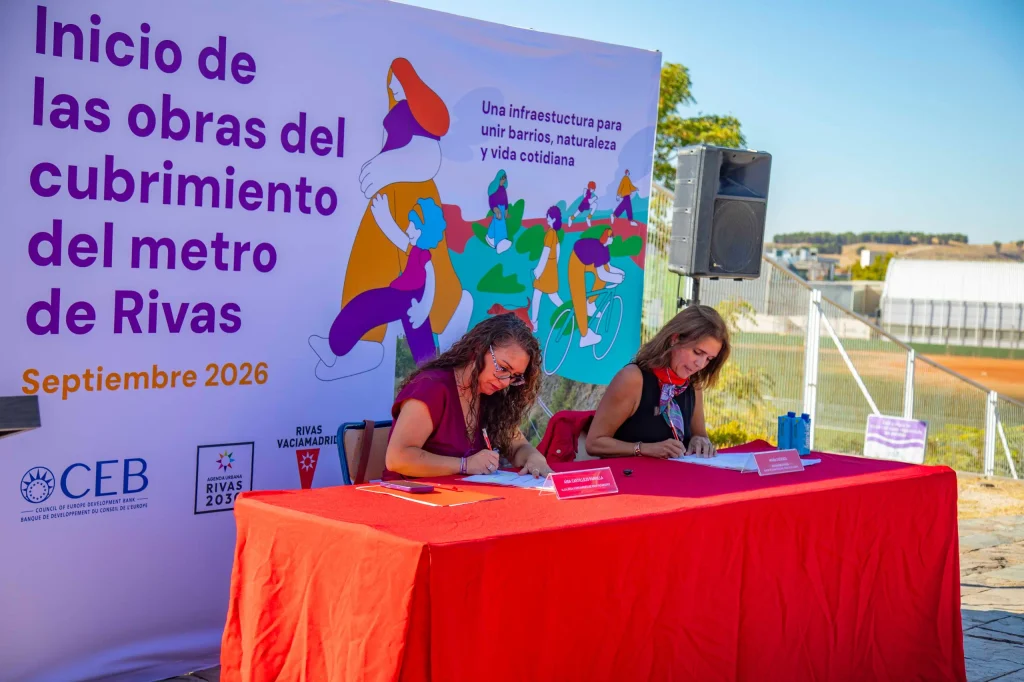 Dos mujeres están sentadas en una mesa roja firmando documentos al aire libre, frente a un gran cartel morado que anuncia el inicio de las obras de cubrimiento de la Línea 9B del Metro en Rivas, con ilustraciones y la fecha de septiembre de 2026.