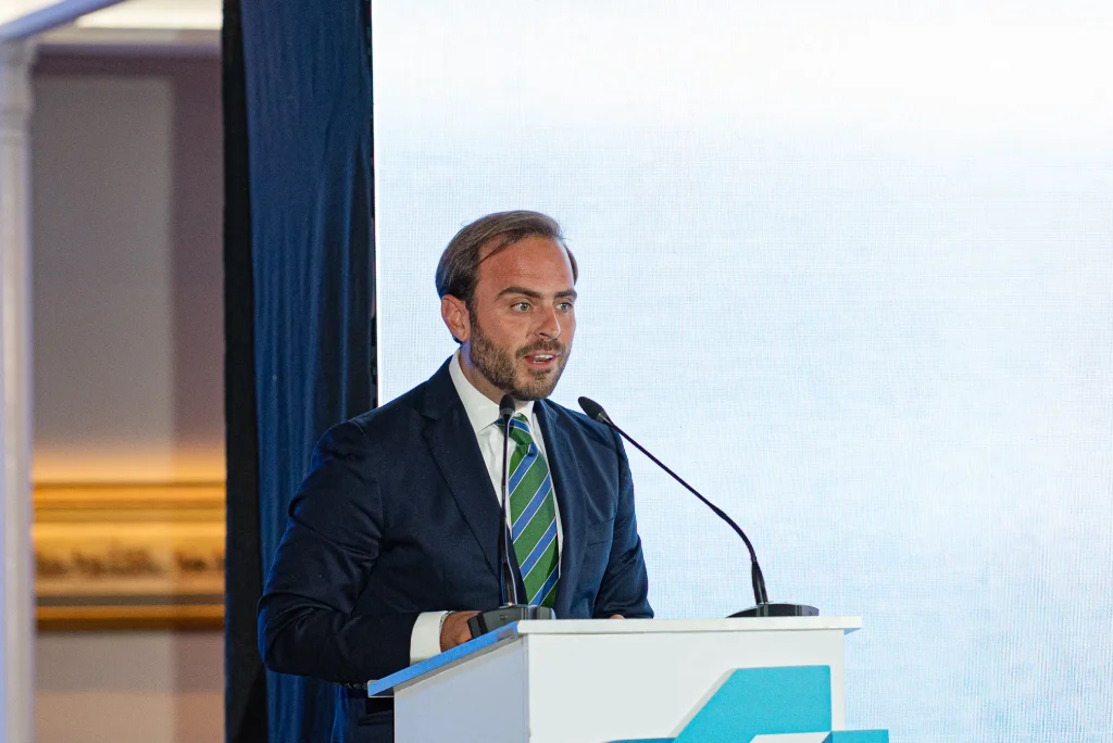 Un hombre vestido con traje y corbata a rayas verdes habla desde un atril con dos micrófonos durante la entrega de los Premios Empresariales de ASEARCO. El fondo es mayoritariamente blanco, con parte de un logotipo azul y una cortina oscura a la izquierda.