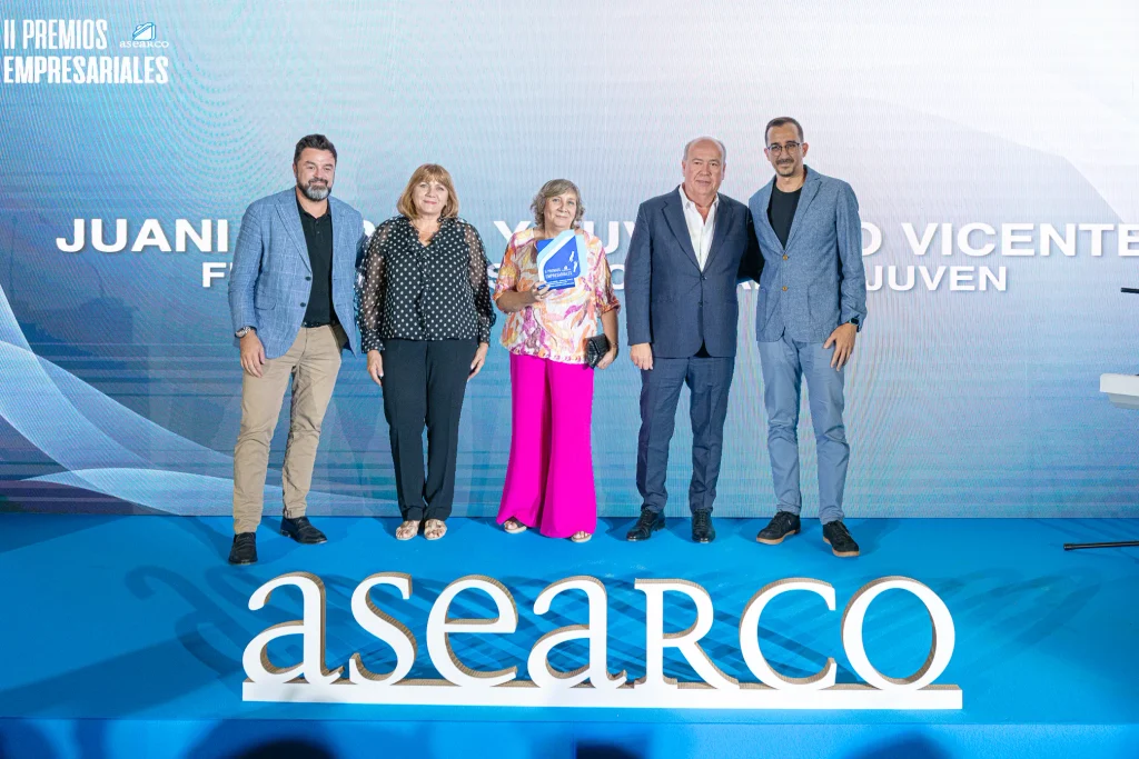 Cinco personas posan en un escenario con un fondo azul; una de ellas sostiene un trofeo. En primer plano aparece la palabra «asearco» escrita en grandes letras blancas. En el telón de fondo se pueden ver los nombres y la inscripción «II Premios Empresariales de ASEARCO».