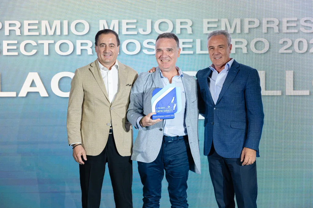 Tres hombres vestidos con traje aparecen sonrientes en el escenario; el que está en el centro sostiene un premio azul y blanco. Detrás de ellos hay un gran cartel en el que se lee: «Premio a la Mejor Empresa del Sector Hostelero 2023». El acto forma parte de los prestigiosos Premios Empresariales de ASEARCO, que reconocen la excelencia en el sector hostelero.
