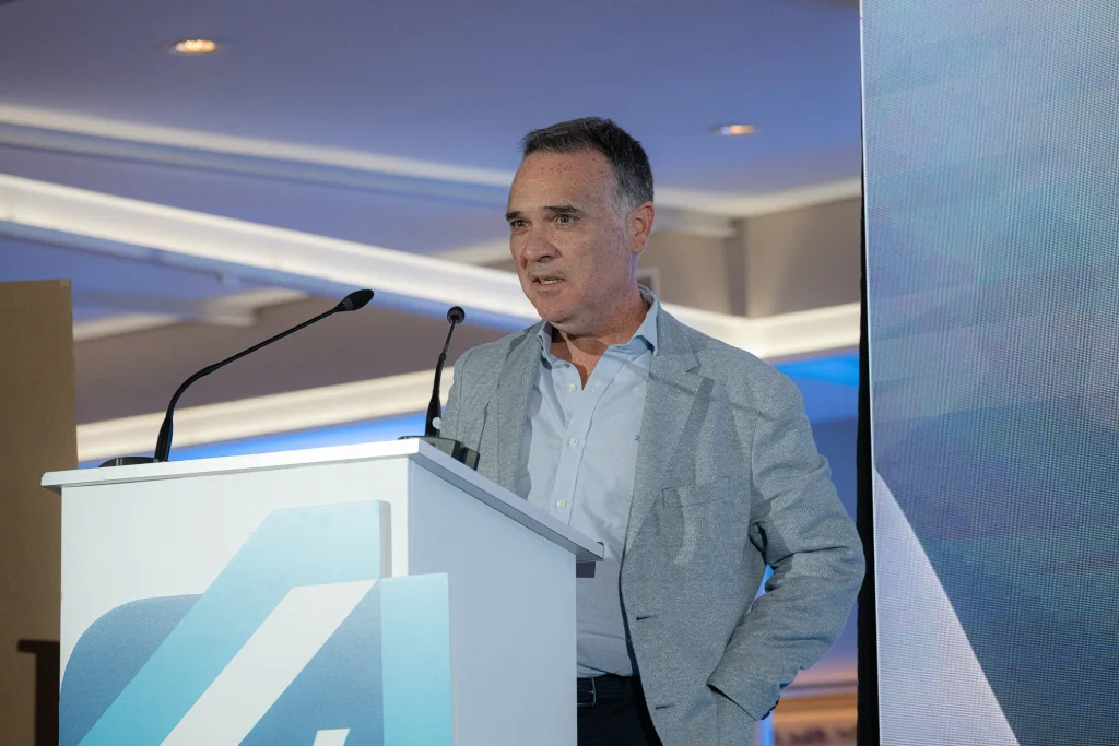 Un hombre con una chaqueta gris claro y una camisa azul está de pie en un atril con dos micrófonos, hablando en un recinto cerrado. El atril luce un logotipo geométrico azul y blanco, que representa los Premios Empresariales de ASEARCO. Al fondo se aprecia una iluminación azul.