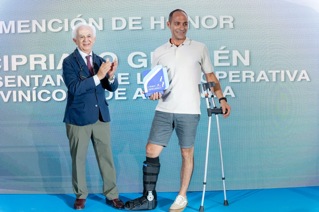 Dos hombres aparecen en el escenario de los Premios Empresariales de ASEARCO; uno está aplaudiendo, mientras que el otro, que lleva una ortesis en la pierna y se apoya en muletas, sostiene un premio y sonríe. Ambos visten ropa semiformal. Sobre un fondo azul se lee un texto en español.