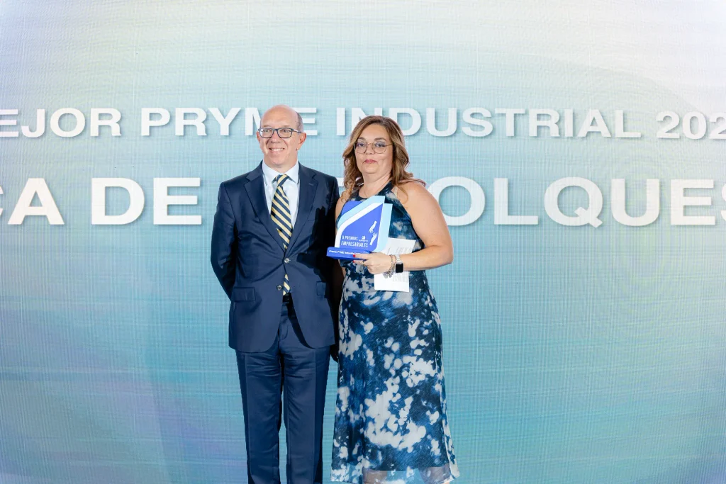 Un hombre vestido con traje y una mujer con un vestido azul estampado sostienen un premio y posan para una foto delante de un telón de fondo con un texto grande que se ve parcialmente, celebrando el reconocimiento recibido en los Premios Empresariales de ASEARCO.