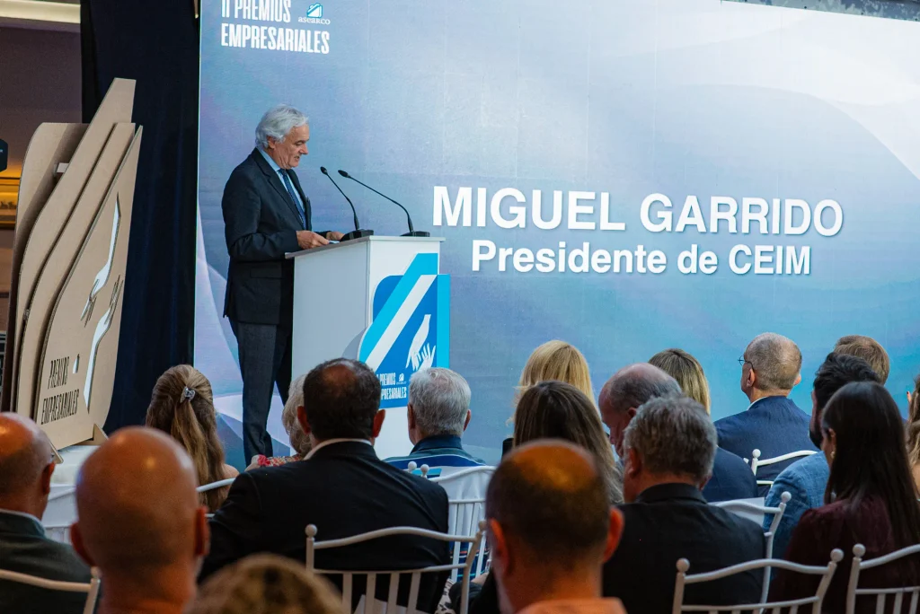 Un hombre se encuentra de pie en un estrado dirigiéndose al público en un acto. En el telón de fondo se lee el texto «Miguel Garrido, presidente de CEIM» y el logotipo de los Premios Empresariales de ASEARCO. El público está sentado y mirando hacia el estrado.