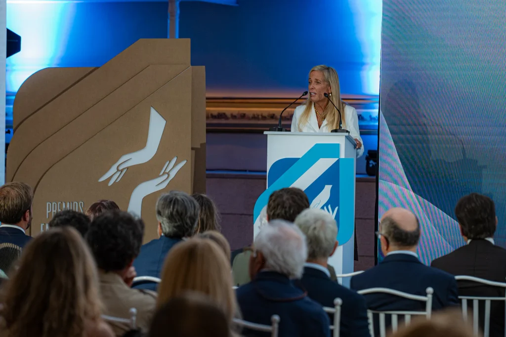 Una mujer se encuentra de pie en un atril dirigiéndose al público en un acto celebrado en un recinto cerrado durante los Premios Empresariales de ASEARCO. El escenario cuenta con iluminación azul y un gran cartel de cartón con dibujos de manos blancas. El público está sentado y mirando hacia la ponente.