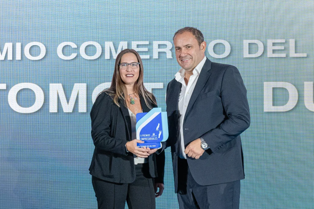 Una mujer y un hombre, vestidos con traje de negocios, posan delante de un cartel con letras grandes. La mujer sostiene un premio azul y blanco de los Premios Empresariales de ASEARCO, y ambos sonríen a la cámara.