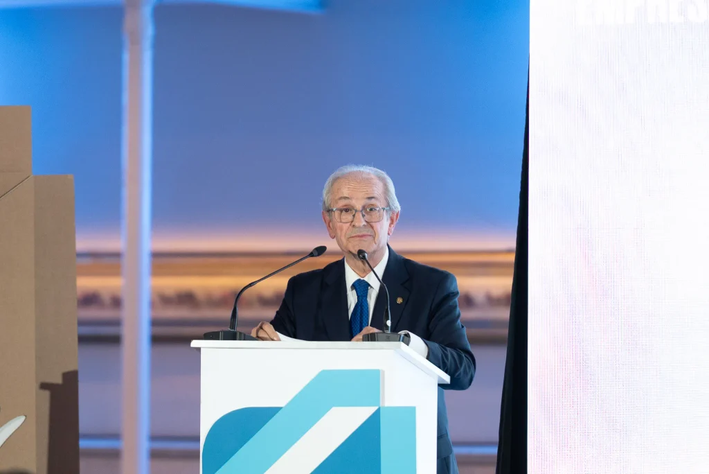 Un hombre mayor, con el pelo canoso y gafas, está de pie en un atril, hablando por dos micrófonos durante la gala de los Premios Empresariales de ASEARCO. El fondo es azul y blanco, y a su derecha se ve una pantalla vertical.