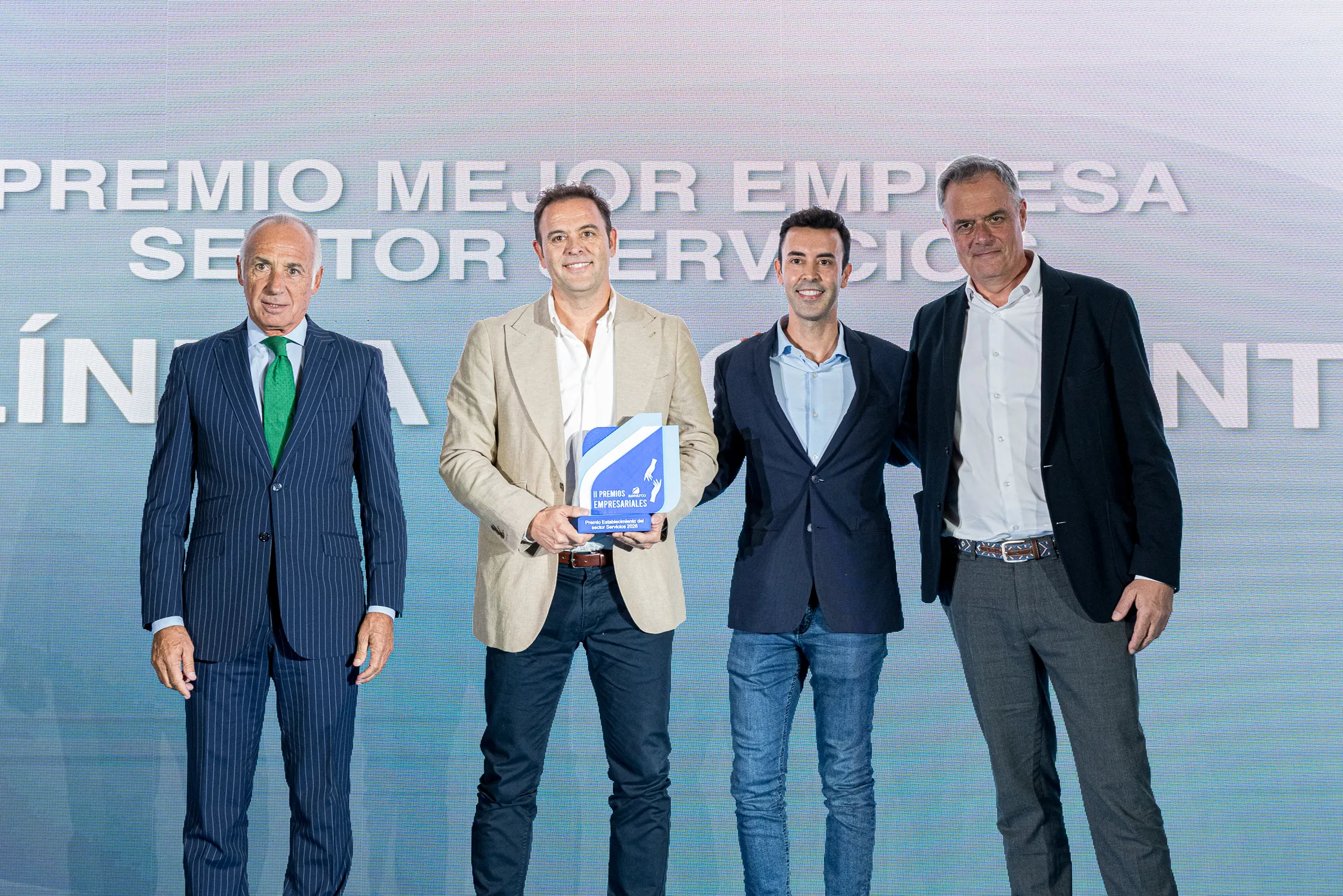 Cuatro hombres vestidos con traje de negocios están de pie en el escenario; uno de ellos sostiene una placa de reconocimiento. Detrás de ellos hay un gran cartel en el que se lee «PREMIO A LA MEJOR EMPRESA DEL SECTOR DE LOS SERVICIOS». El grupo posa para una foto formal en la gala de los Premios Empresariales de ASEARCO.