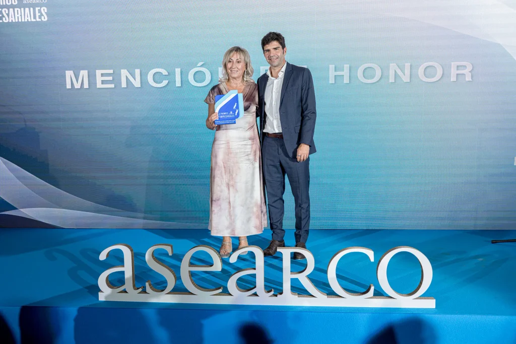 Una mujer que sostiene un premio aparece de pie junto a un hombre en un escenario azul. Detrás de ellos, en una pantalla se lee «MENCIÓN DE HONOR» y, frente a ellos, hay un gran cartel de ASEARCO que destaca la importancia de los Premios Empresariales de ASEARCO. Ambos van vestidos de forma elegante.