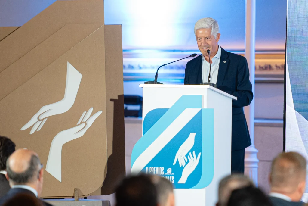 Un hombre mayor, vestido con traje, se encuentra de pie en un atril con un micrófono, dirigiéndose al público durante los Premios Empresariales de ASEARCO. El atril y el telón de fondo presentan ilustraciones estilizadas de manos. En primer plano hay varias personas sentadas, mirando hacia el orador.