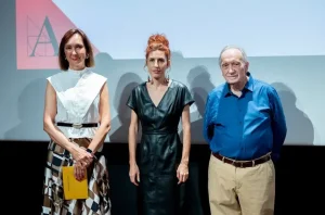Tres adultos, que participan en el evento «Óscar España» de preselección, posan delante de una pantalla de proyección. La mujer de la izquierda lleva un top blanco y una falda estampada; la mujer del centro, un vestido negro; y el hombre de la derecha, una camisa azul y unos pantalones beige.