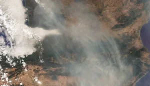 Una imagen de satélite muestra el humo de los incendios forestales en España extendiéndose por un paisaje seco, con nubes a la izquierda y elementos del terreno visibles a través de la neblina. En la esquina superior derecha se aprecian la costa y una gran masa de agua.