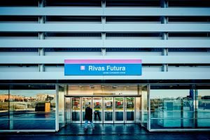 Entrada de la estación de Rivas Futura: bajo el cartel azul con la banda rosa y el logotipo de Metro de Madrid, la puerta acristalada de la estación y una persona caminando, en una imagen nocturna.